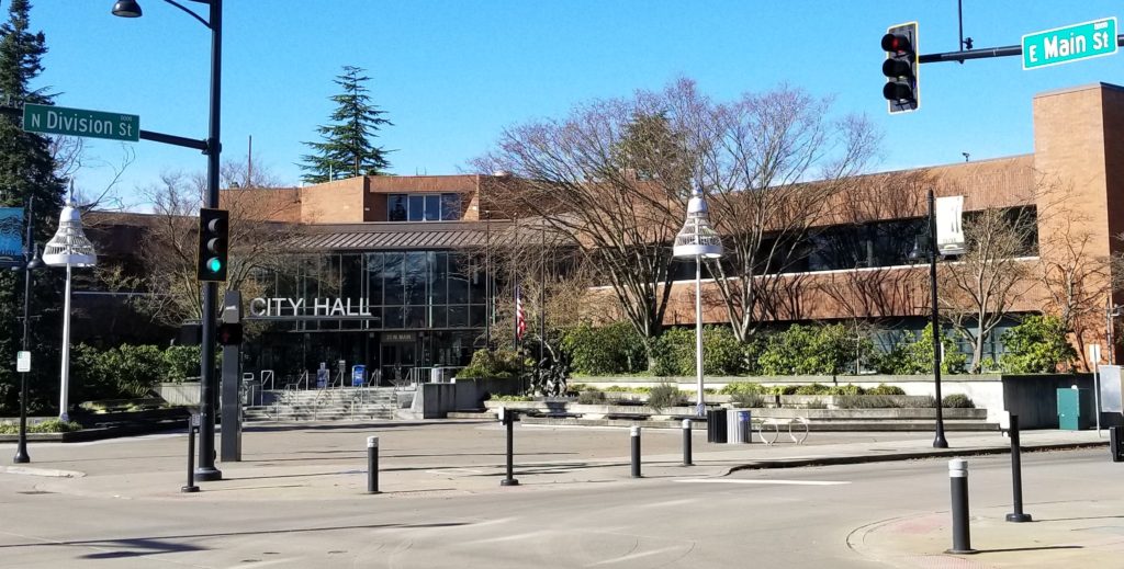 Auburn City Hall, a brick building with dark glass windows and doors throughout. Trees and greenery surround the building, with tall silver bell shaped lamps in the court yard in front of the building. The photo is taken from across the street at an angle, capturing a portion of the street and street lights.