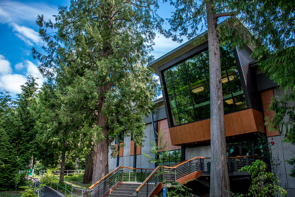 Cedar Hall Green River College, a glass front building with an angled front, sloping down, on the second story. Large evergreen trees surround the building.