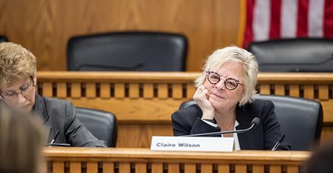senator Claire Wilson rests her chin on her hand, her head tilting slightly to the side as she listens to someone speak during a committee meeting, a subtle smile on her face