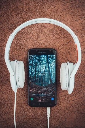 A set of white headphones is set above a smartphone, a forest shown on the screen. The ears of the headphones are placed on either side of the phone.