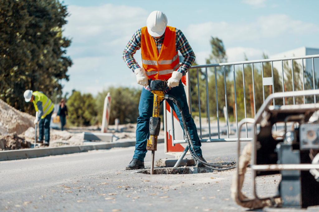 A male construction worker wearing an orange safety vest and white hardhat uses a jackhammer on an asphalt sidewalk