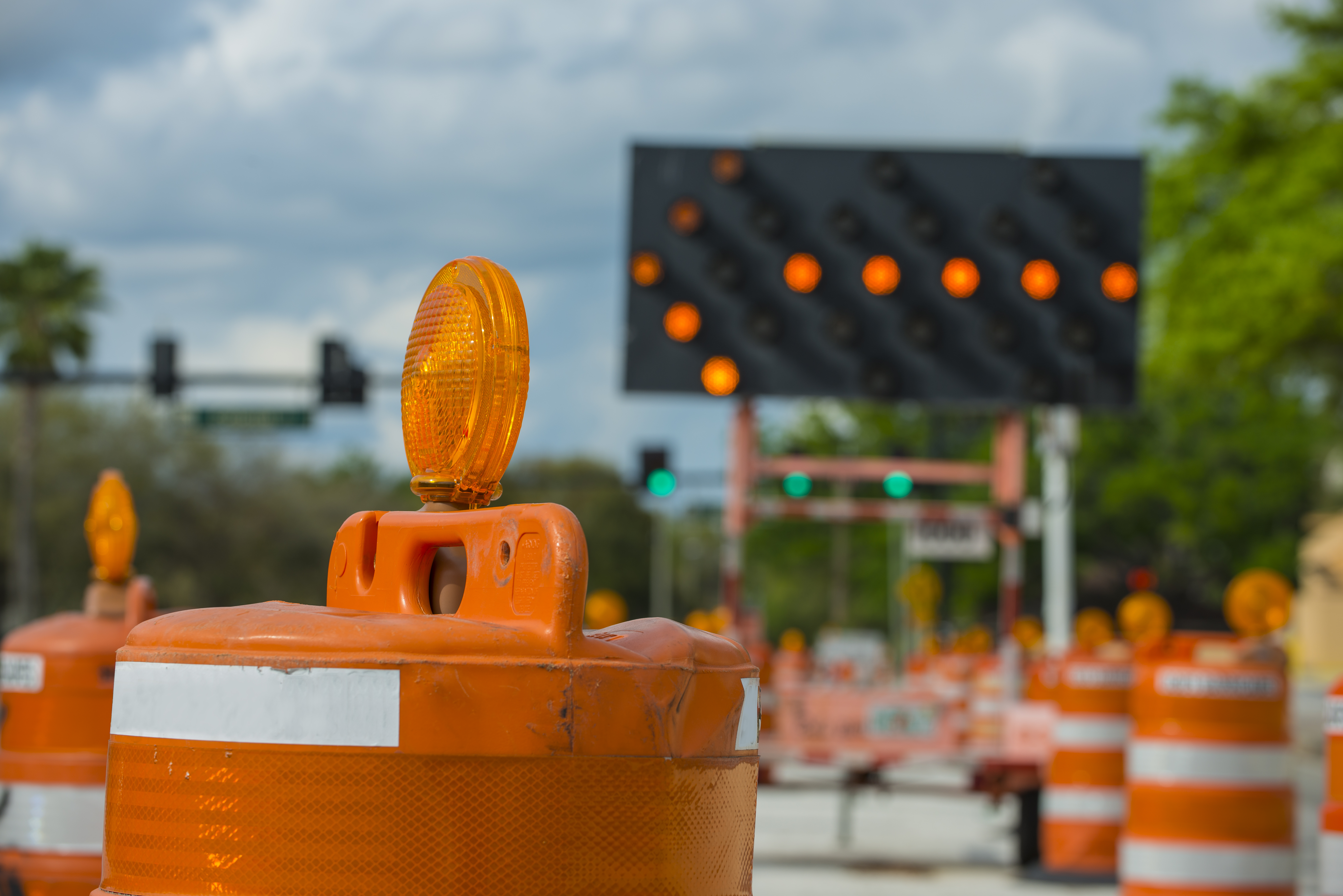 Traffic barrels and cones surround a road construction site with a large electric sign with an arrow pointing left in the background