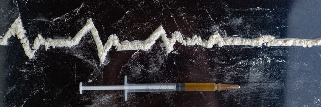a needle with dark yellow liquid in it lays beneither a heartbeat line that flattens, the line is made of white powder - representing an overdose