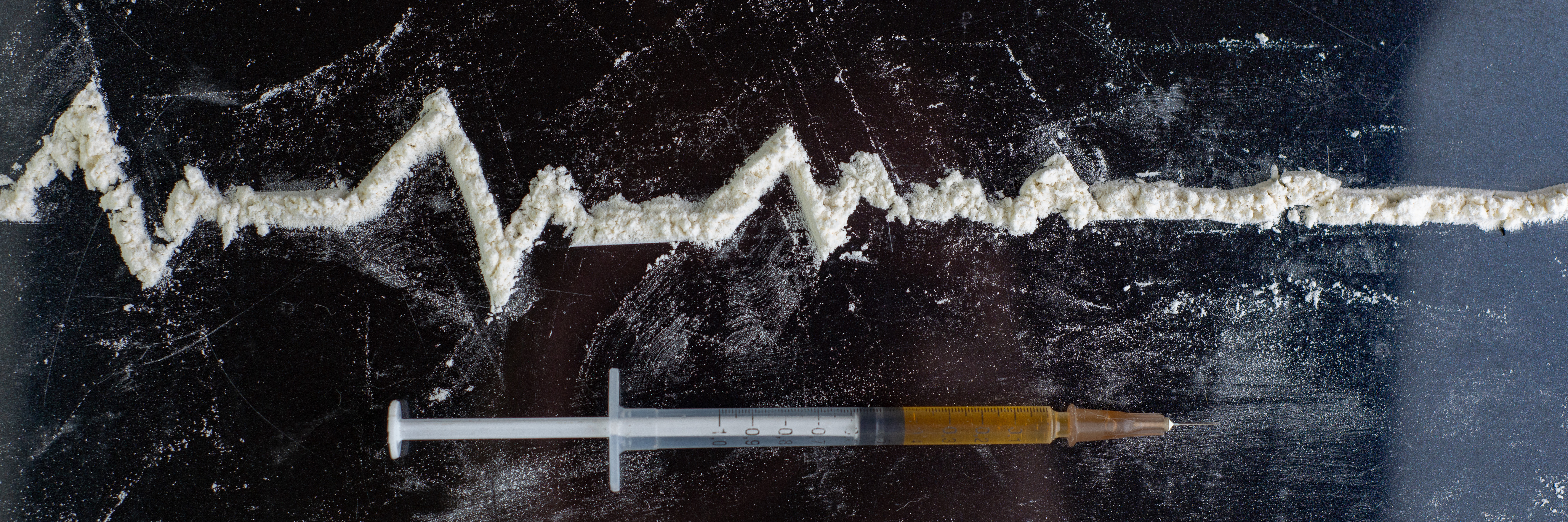 a needle with dark yellow liquid in it lays beneither a heartbeat line that flattens, the line is made of white powder - representing an overdose