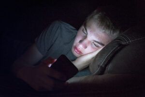 a teen lays down in the dark staring at his brightly lit smart phone