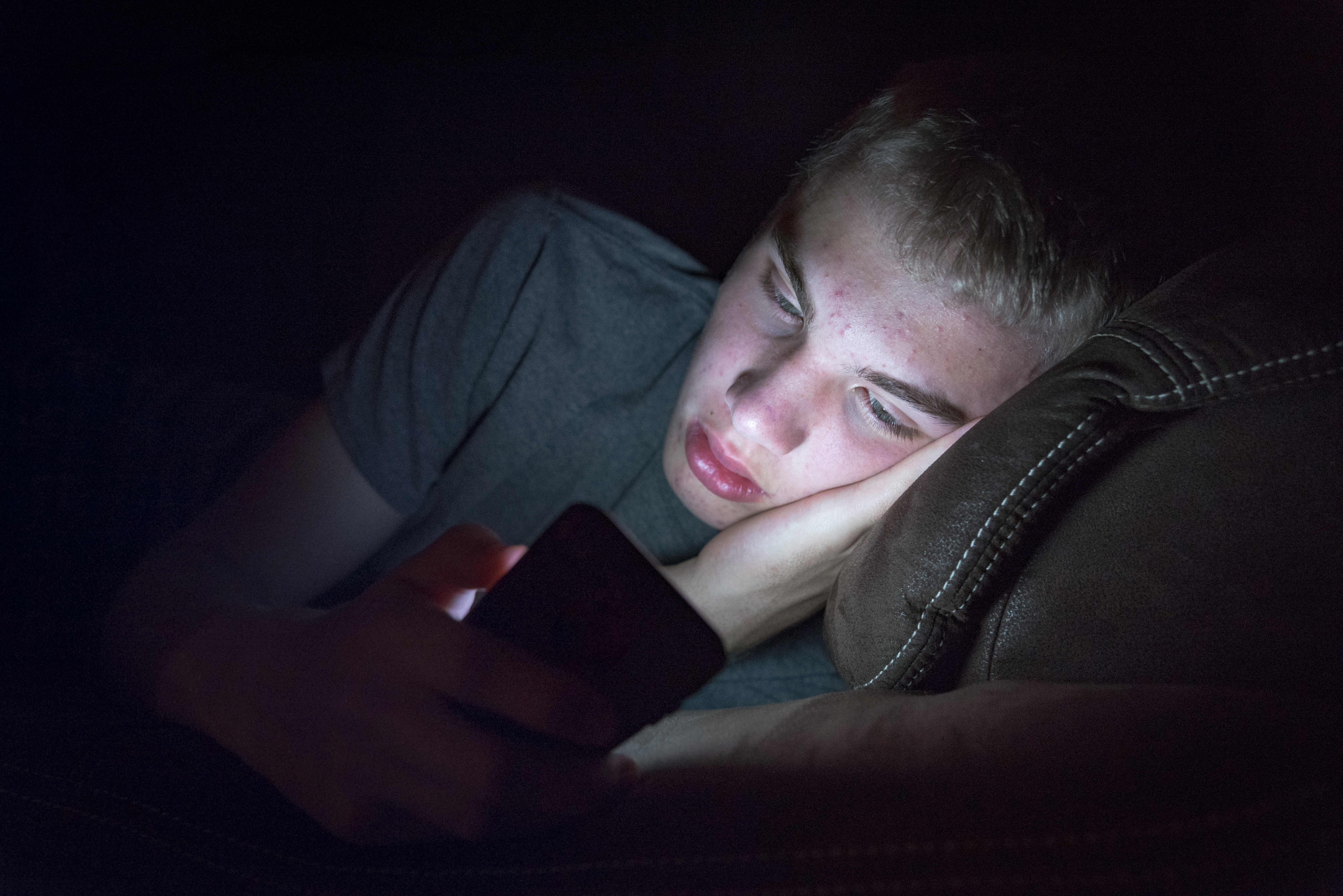 a teen lays down in the dark staring at his brightly lit smart phone