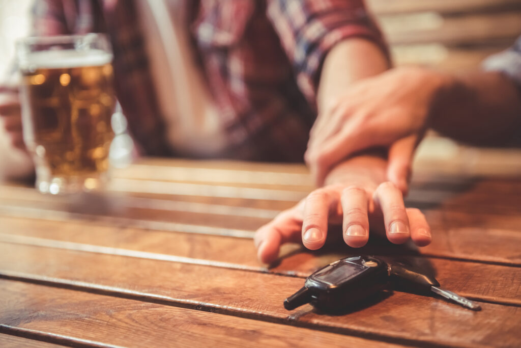 A male reaches for a set of keys on a bar, a beer in his other hand. Another person holds his forearm in protest.