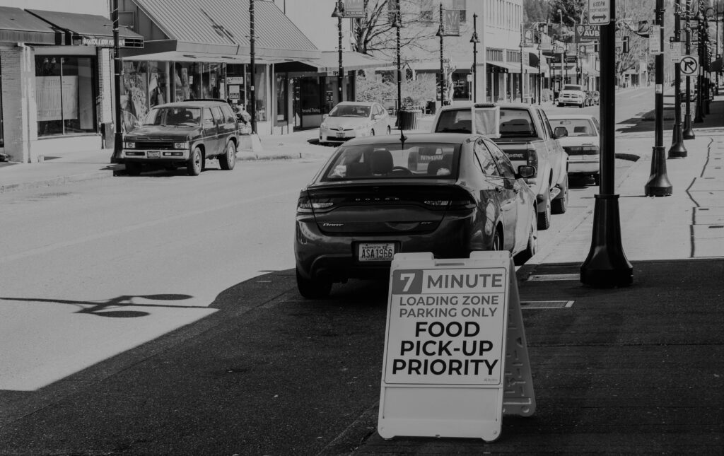 A black and white photo of downtown E Main Street. The photo is taken from the sidewalk looking up toward Auburn Way South. On the sidewalk is a sign stating "Food Pick Up Priority"
