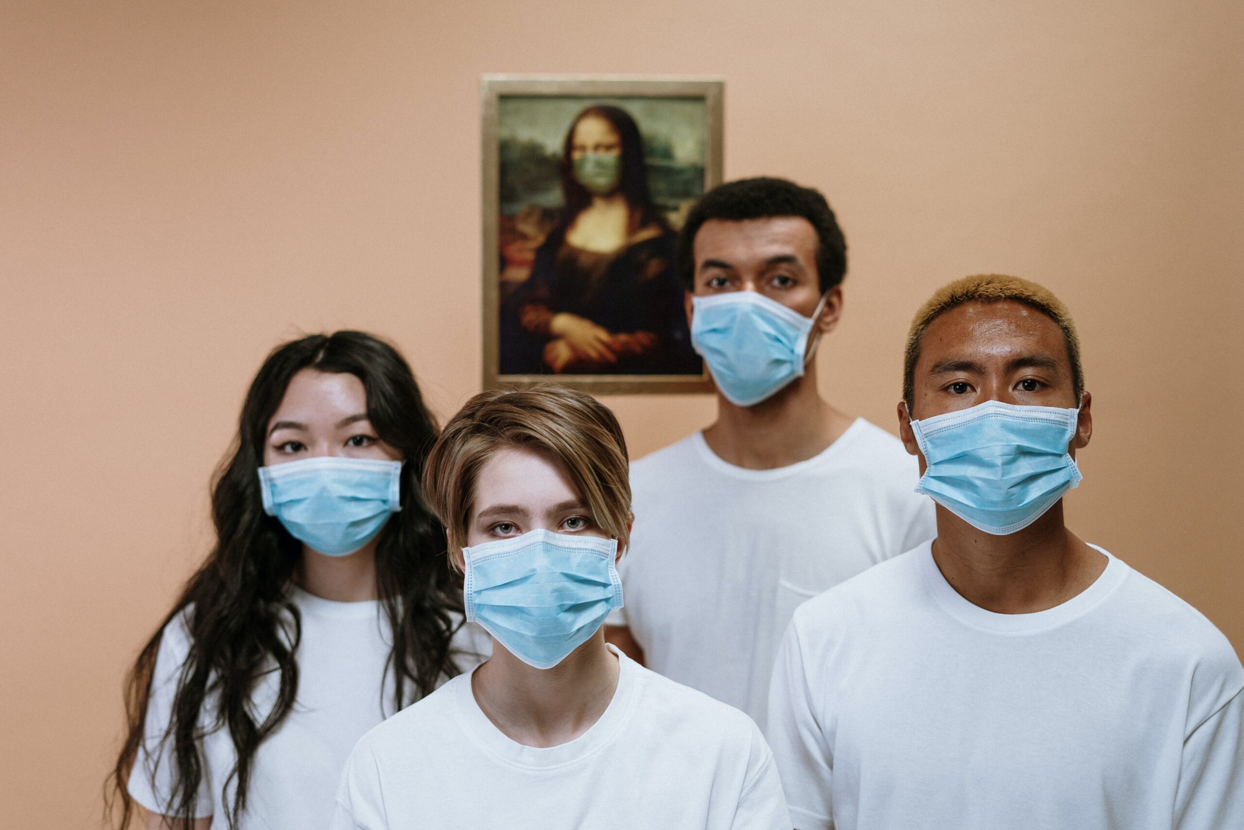 A group of diverse individuals, 2 females and 2 males, stand together weaing white shirts and blue surgical masks. Behind them on a tan wall is a Mona Lisa painting, also wearing a mask.
