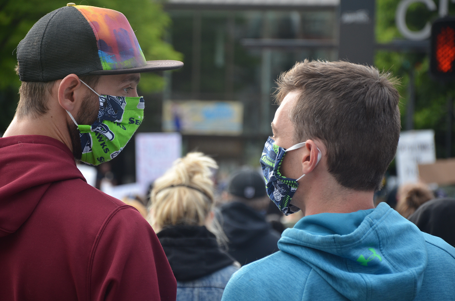 two adult males wearing face masks and hoodies look at each other during a large gathering outside of city hall.