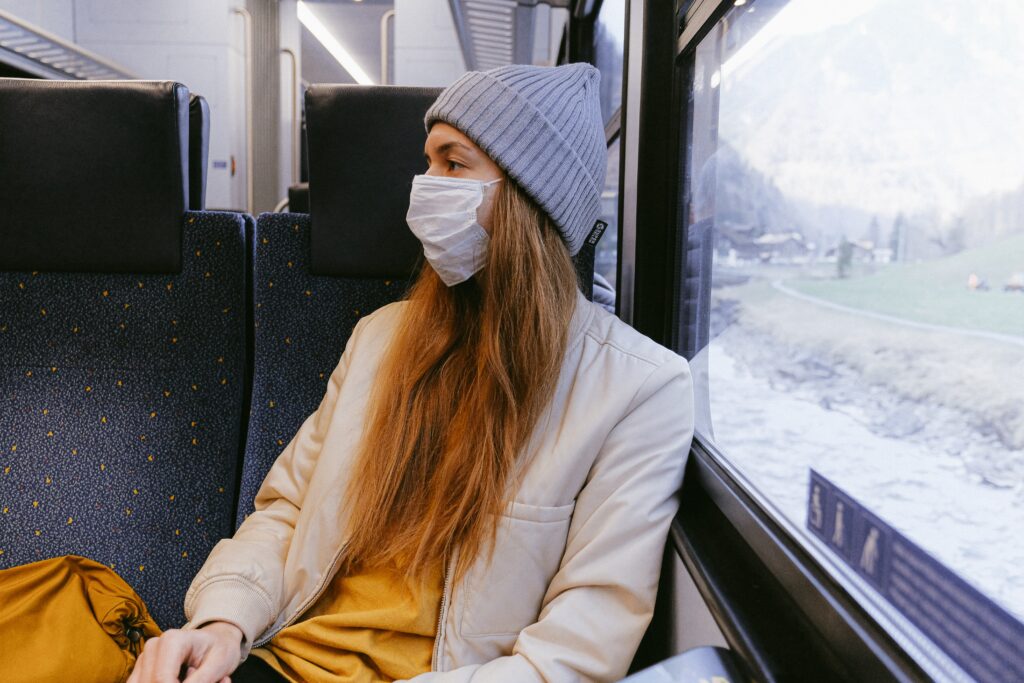 woman-in-gray-knit-cap-and-beige-coat-wearing a masks sits on a train with her back toward the window.