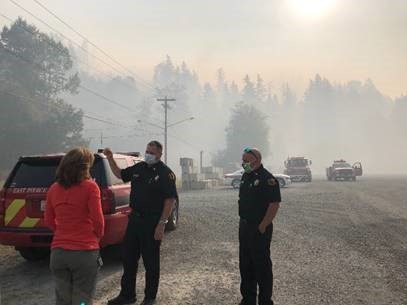 Two fire professionals speak with Rep. Schrier next to a fire pick up truck, the air around them covered in a hazy blanket of smoke
