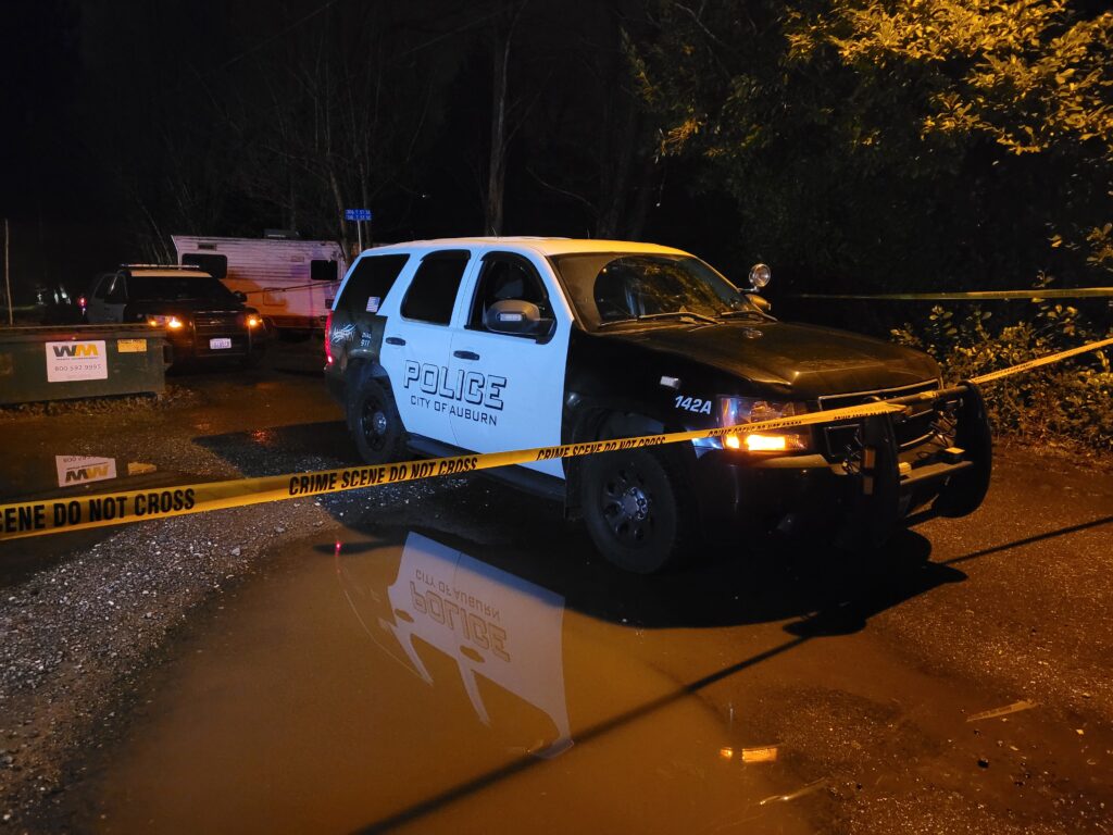 An Auburn Police Department SUV is parked on a wet gravely road, a large muddy puddle next to the vehicle reflects the side of the SUV, including the APD logo. Yellow crime scene tape crosses over the front of the SUV and along far bushes in the background. Behind the SUV is a second APD suv, a white trailer is parked at an angle to the side of that vehicle.