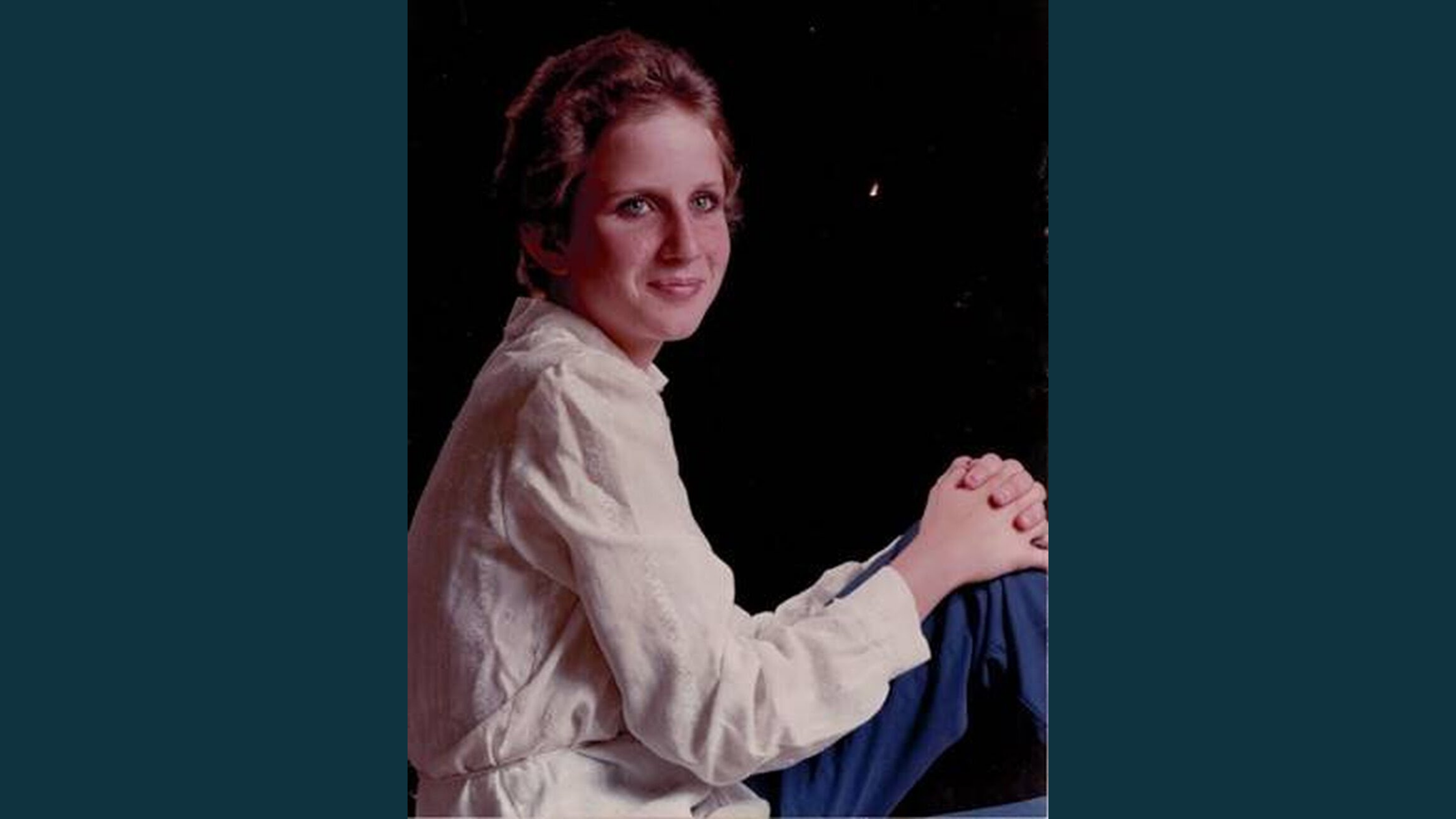 A photo of Wendy Stephens, a young teenage white female with short dark blonde hair. She sits with her body facing to the right, one knee bent up with her hands clasped around it. She looks toward the camera with a soft smile. Wendy wears a pale pink long sleeve shirt and dark blue jeans. She is set against a black background.