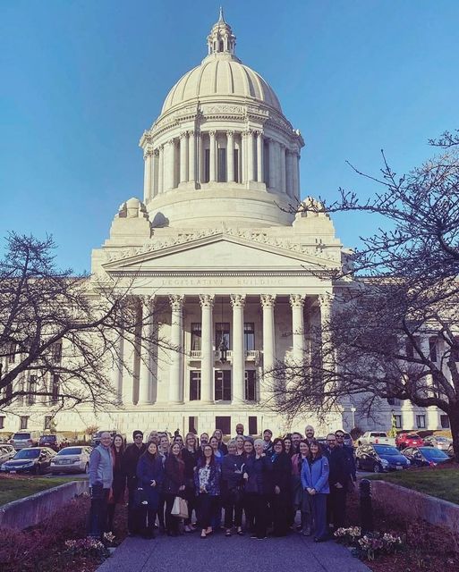 A group of individuals stand together on the sidewalk between two short walls, trees without leaves on either side. In the background a large domed building, the Washington State Capitol building in Olympia.. 