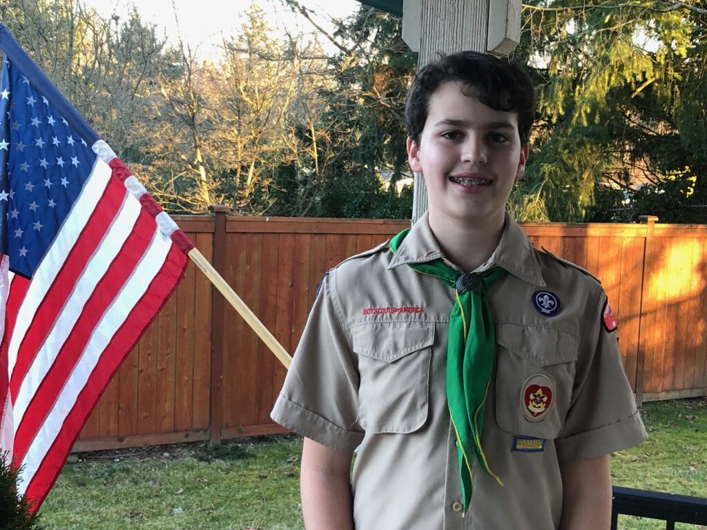 Auburn Boy Scout Alec Mayer, a white male teenager with short brown hair and dark eyes, stands on a porch in a back yard. He wears a typical tan Boy Scout uniform and green kerchief, with two round patches on the right side of his chest and 'Boy Scouts of America' over the left pocket of his shirt. An American flag hangs from the porch post to the left of Alec.