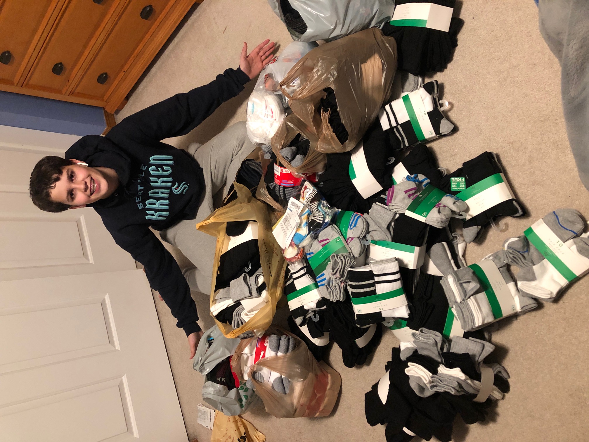 Alec Mayer, an Auburn Boy Scout, sits on the floor of a bedroom, his arms outstretched showing the large pile of black, white, and gray socks spilling out of his lap. Alec, a white teenage male with short brown hair, wears a Seattle Kraken NHL sweatshirt and gray pants.
