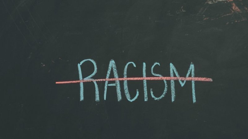 The word "racism" is written in light green chalk on a dark green chalk board and is crossed out with a red line.
