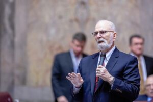 Senator Phil Fortunato, an older white male with white hair and a trimmed white beard, holds a microphone as he speaks on the State Senate floor. He is dressed in a dark blue suit. Two men in suits are seen in the background against the tan marble wall, their features out of focus.