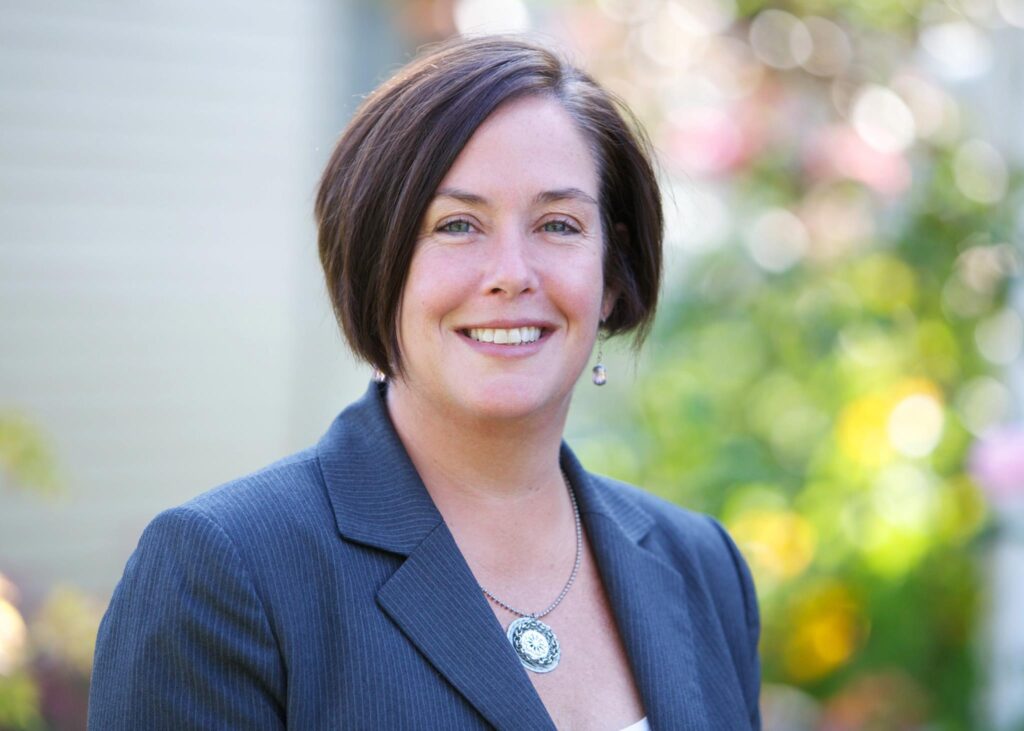 Stephanie bowman, a a brunette Caucasian female, looks toward the camera while standing slightly angle to the left. She wears a blue blazer and silver necklace with circle pendant. The background is blurred landscaping.