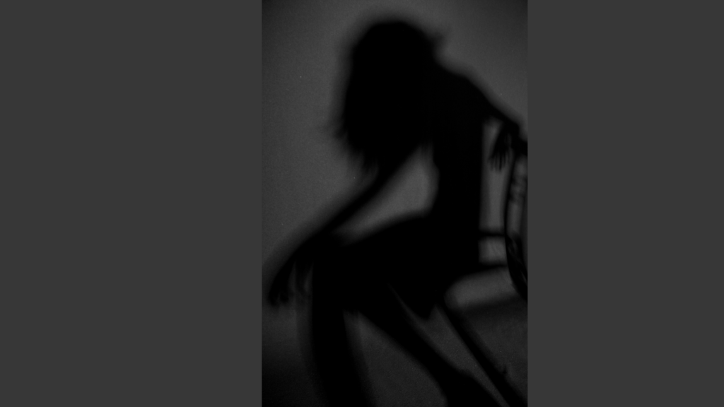 A shadow figure of a female sits forward and slightly hunched over on a chair. The background is differing hues of medium to dark gray.