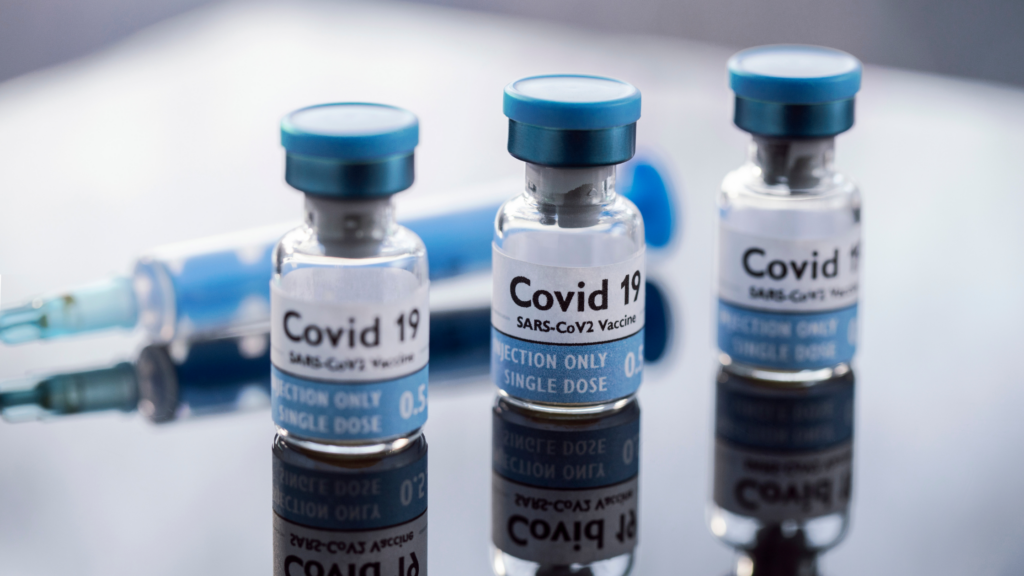 Three small bottles of the COVID-19 vaccine. Behind the three bottles lays a single syringe.