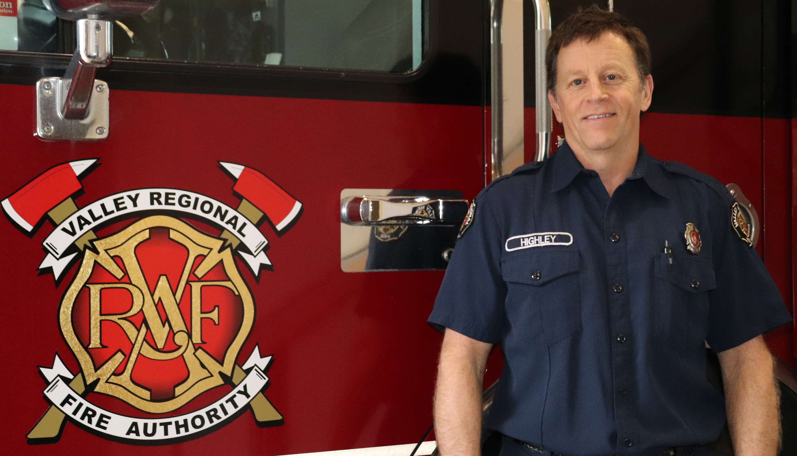 A photo of Valley Regional Fire Authority Firefighter Will Highley, a white middle-aged male with short brown hair wearing a dark navy VRFA uniform shirt with his name on the left side of his chest. He smiles as he stands in front of a VRFA fire truck.