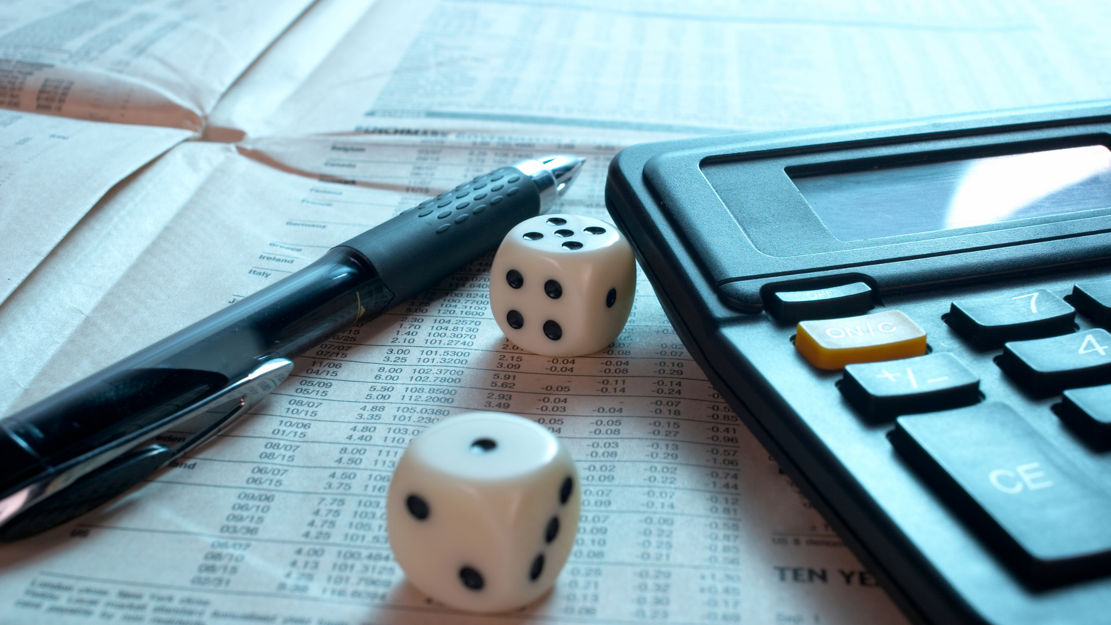 A pen, pair of white and black dice, and manual calculator sit on an open newspaper. The paper is open to a stock trading page.