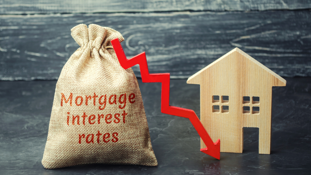 A burlap sack sack with the words "mortgage interest rates" sits to the left of a wooden block in the shape of a house. A red line with an arrow on the end starts at the top of the sack and points down toward the based of the house, with sharp ups and downs in it as it falls. The photo's background is a faded black and off-white wood panel.