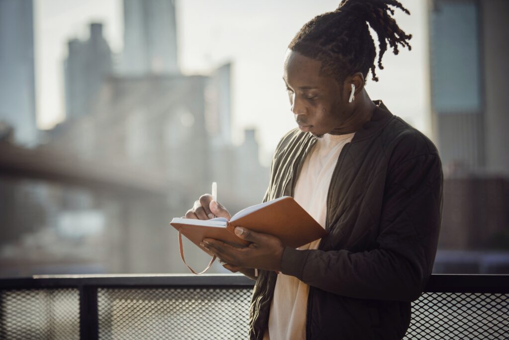 A twenty something Black male with dreads pulled into a high pony tail stands on a balcony writing in a tan journal. White air pods rest in his ears as he writes.