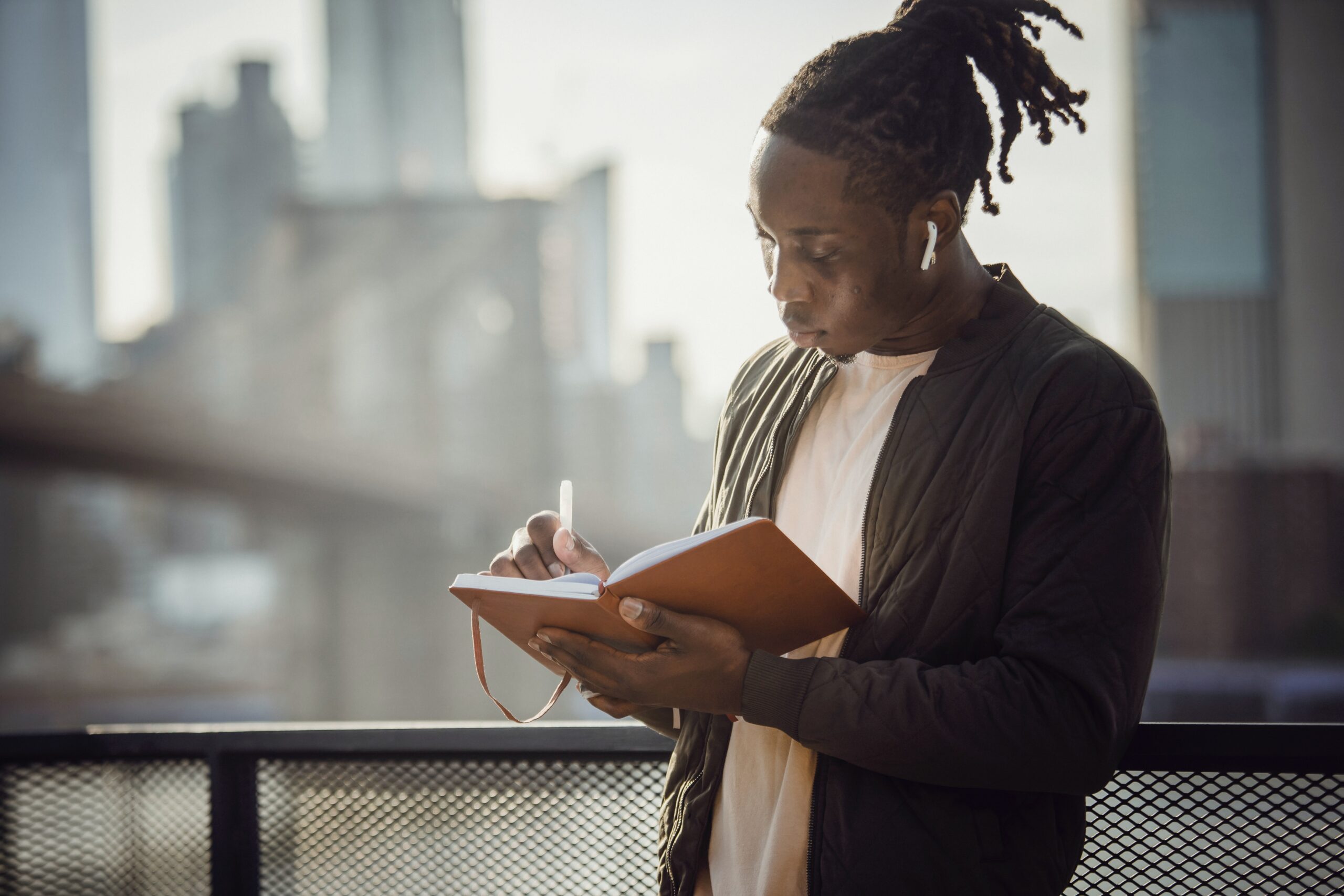A twenty something Black male with dreads pulled into a high pony tail stands on a balcony writing in a tan journal. White air pods rest in his ears as he writes.