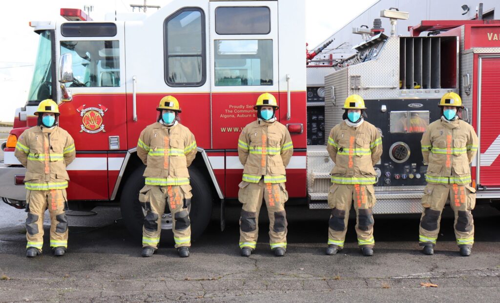 5 firefighters in full gear, wearing surgical masks, stand in front of a of the side of a red and white fire engine 