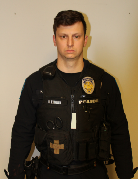 A white male with short brown hair stands against a wall looking forward. He wears an Auburn Police Uniform with the name patch "K Lyman." His firearm is not in its holster and there is a 