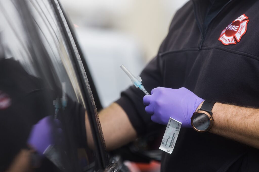 A hand wearing purple gloves holds a needle with the COVID-19 vaccine, a Band-Aid stuck to the back of their hand, the other hand reaches into an open car window.