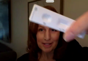 A white female, Congresswoman Kim Schrier, holds up a rectangular test. The flat white plastic test has an open oval space and an rectangle slot on it's front.