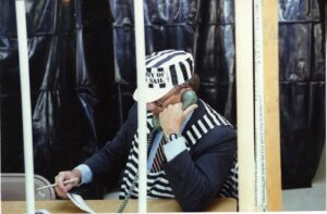 A man sits at a desk behind bars. He is ressed in a jail inmate costume. The man is on the phone looking at a piece of paper in his other hand
