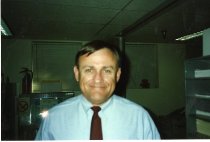 A white male with dark blonde hair in a light blue dress shirt and dark red tie looks at the camera with a closed mouth smile.
