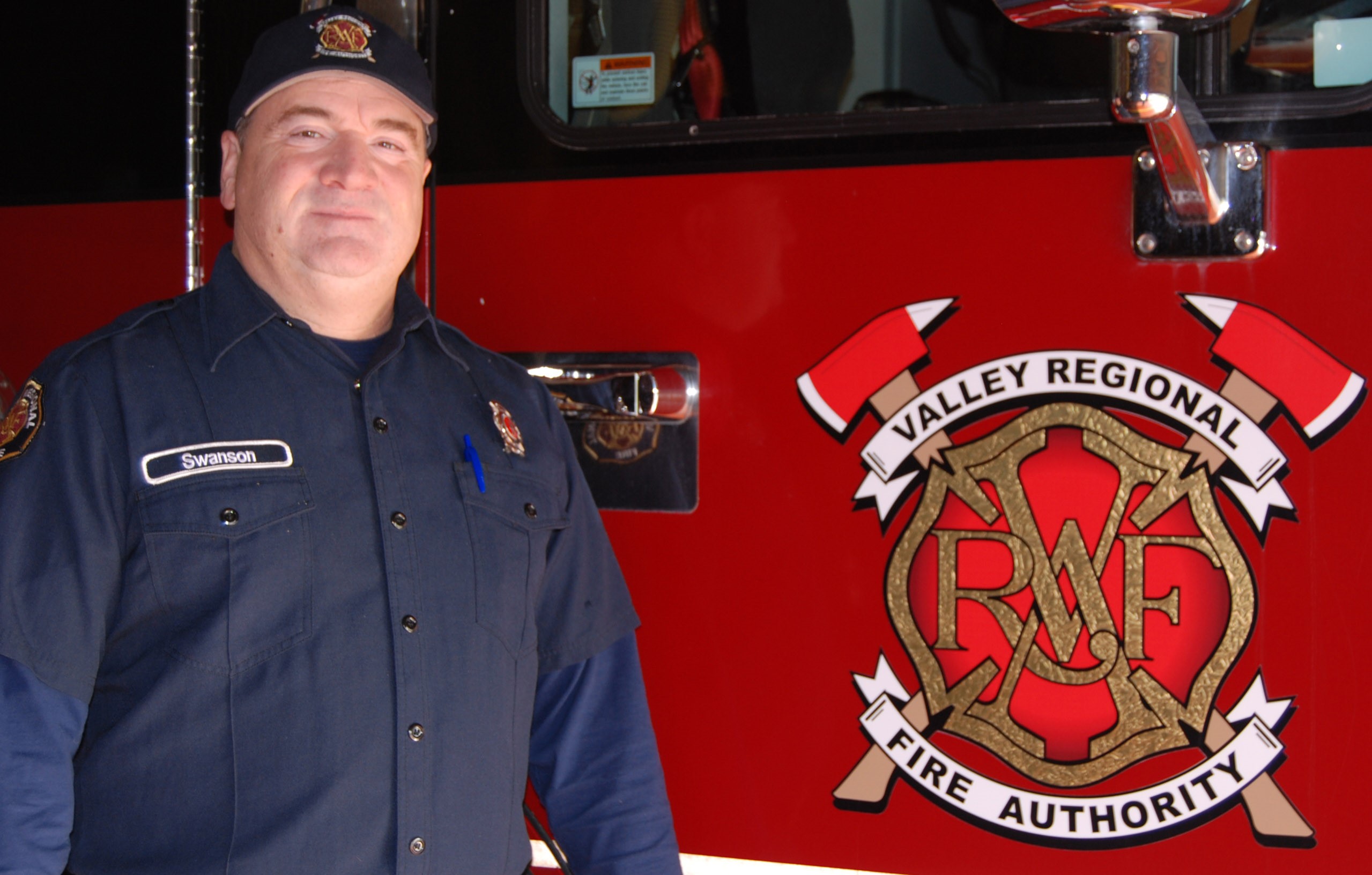A male firefighter in a dark navy uniform stands in front of a fire engine, the VRFA logo shown on the truck's door.