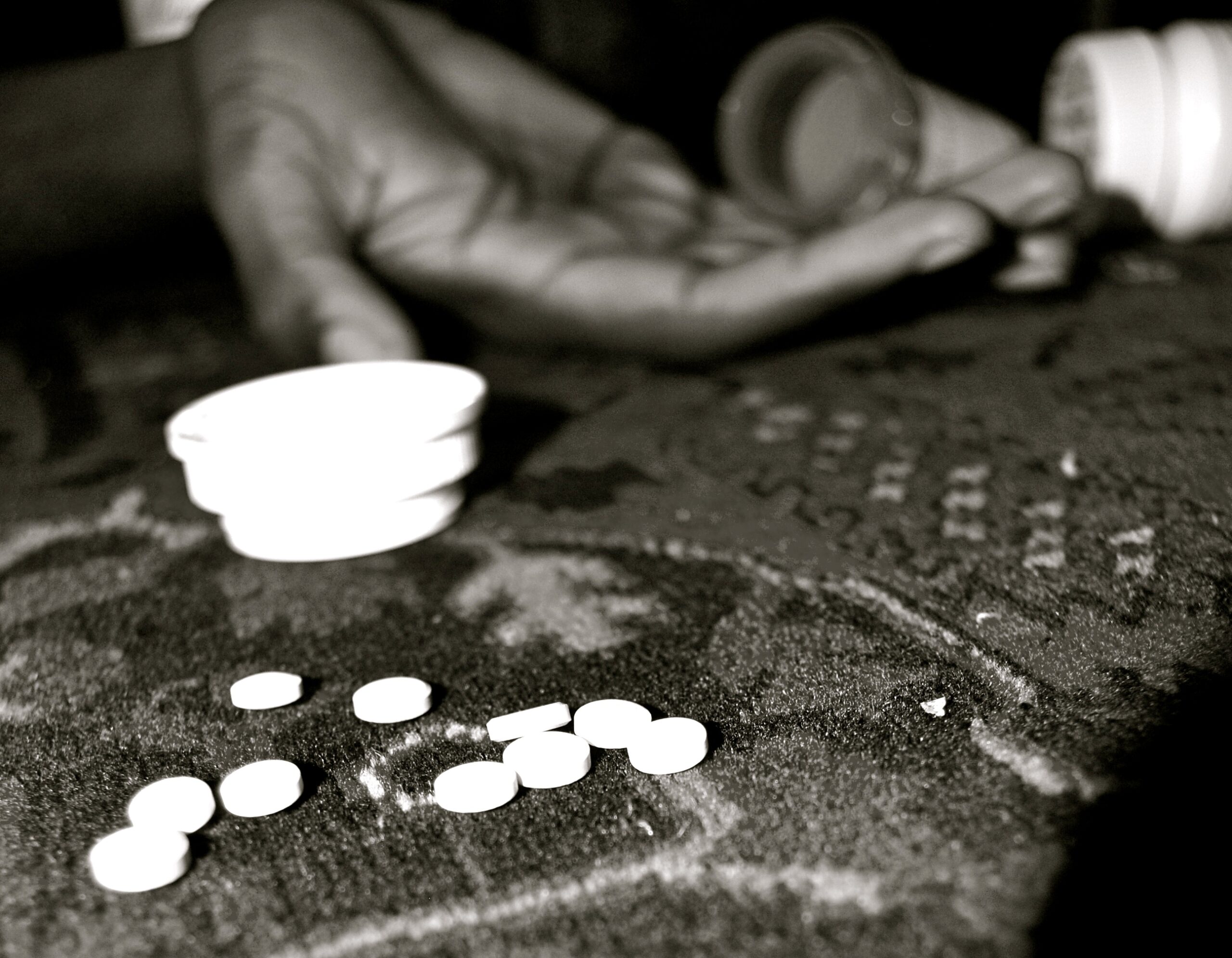 A cluster of white pills is focused in the foreground, a white pill bottle top is flipped on its side, a hand rests in the background, cupped with two pill bottles resting on its edge poised to pour.