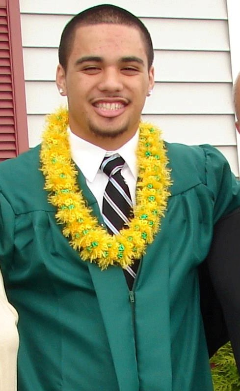 A young man in a green graduation robe with a striped tie and bright yellow lei smiles at the camera