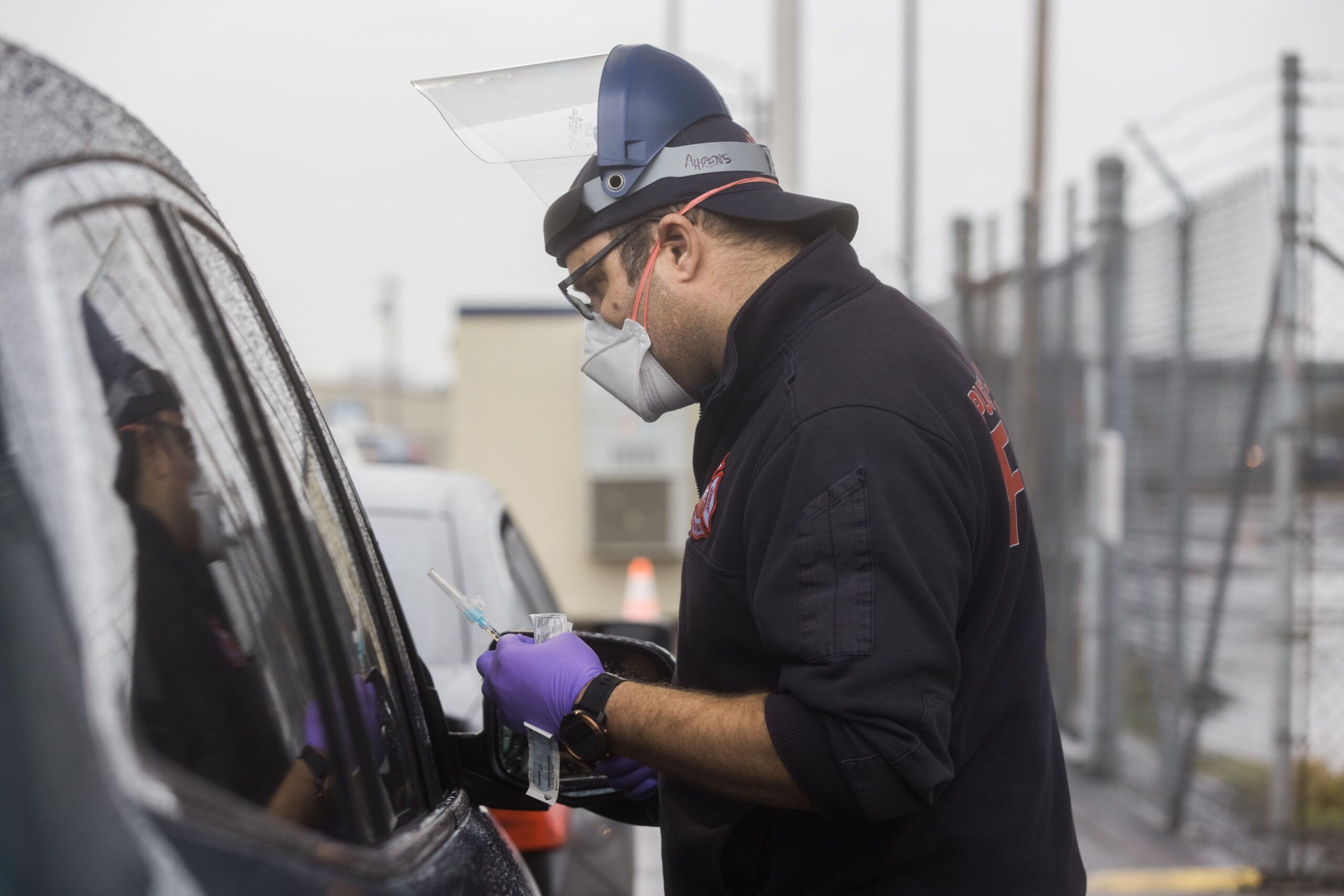 A male wearing blue surgical mask, clear plastic face shield and black coat looks into the driver's window of a vehicle while holding a syringe and needle, a bandaid stuck to the back of his hand. he male reaching in to the driver, whose face is largely obscured by a wet windshield, indicates he is administering a shot.