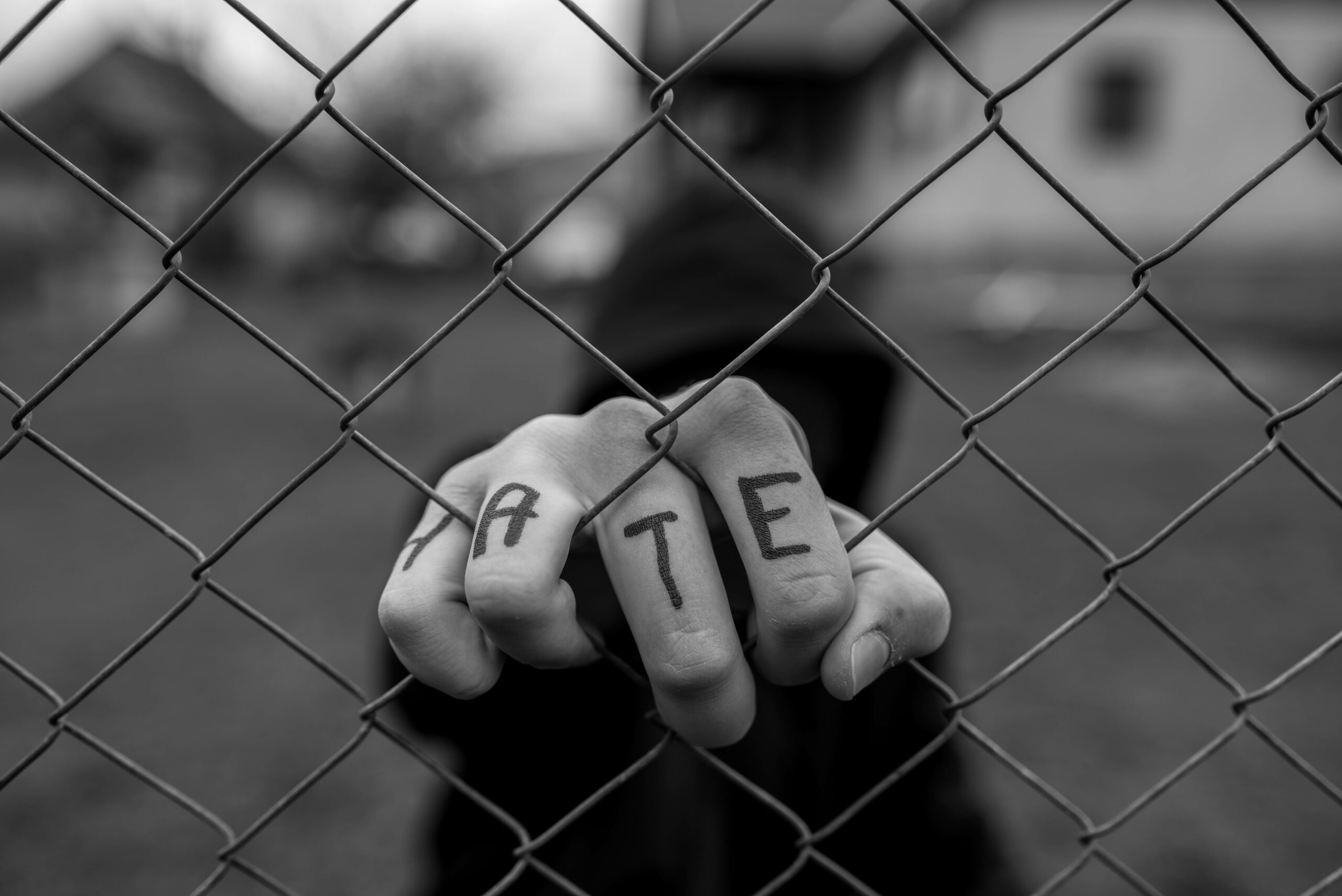 A black and white photo of a hand grasping a wire fence. H A T E is written on the four fingers pulling through the fence.