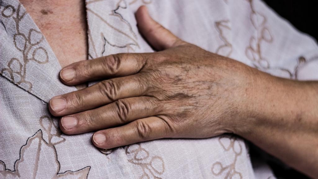 The hand of an elderly Black woman rests on her chest, over her heart. She wears a pale brown shirt with a tan design.