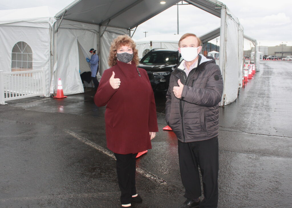 A middle-aged woman with light brown hair wearing a cloth mask and maroon coat stands next to senior man in a white mask and dark gray coat.  Each presents a thumbs up as they stand in front of a large tent with a car under it.