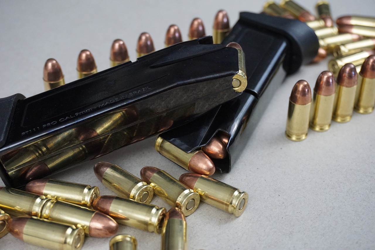 Two black, loaded, pistol magazines lay on their sides surrounded by a handful of loose bullets. The gold and copper unspent bullets in varying positions from from standing in a straight line, and laying in a pile.