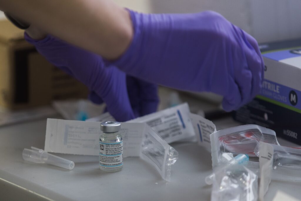 Hands wearing purple gloves hover above opened packages of needles, a small clear bottle with liquid inside among the wrappings. It appears the individuals is preparing vaccines for use.