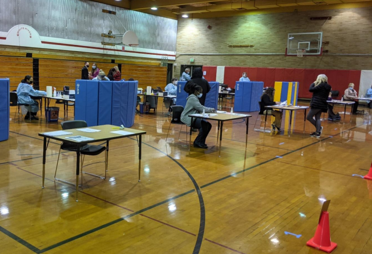 Individual testing stations (tables and chairs behind gym mats stood on end) are set up in a school gym. Individuals are throughout the gym at the stations, standing speaking, and checking in.