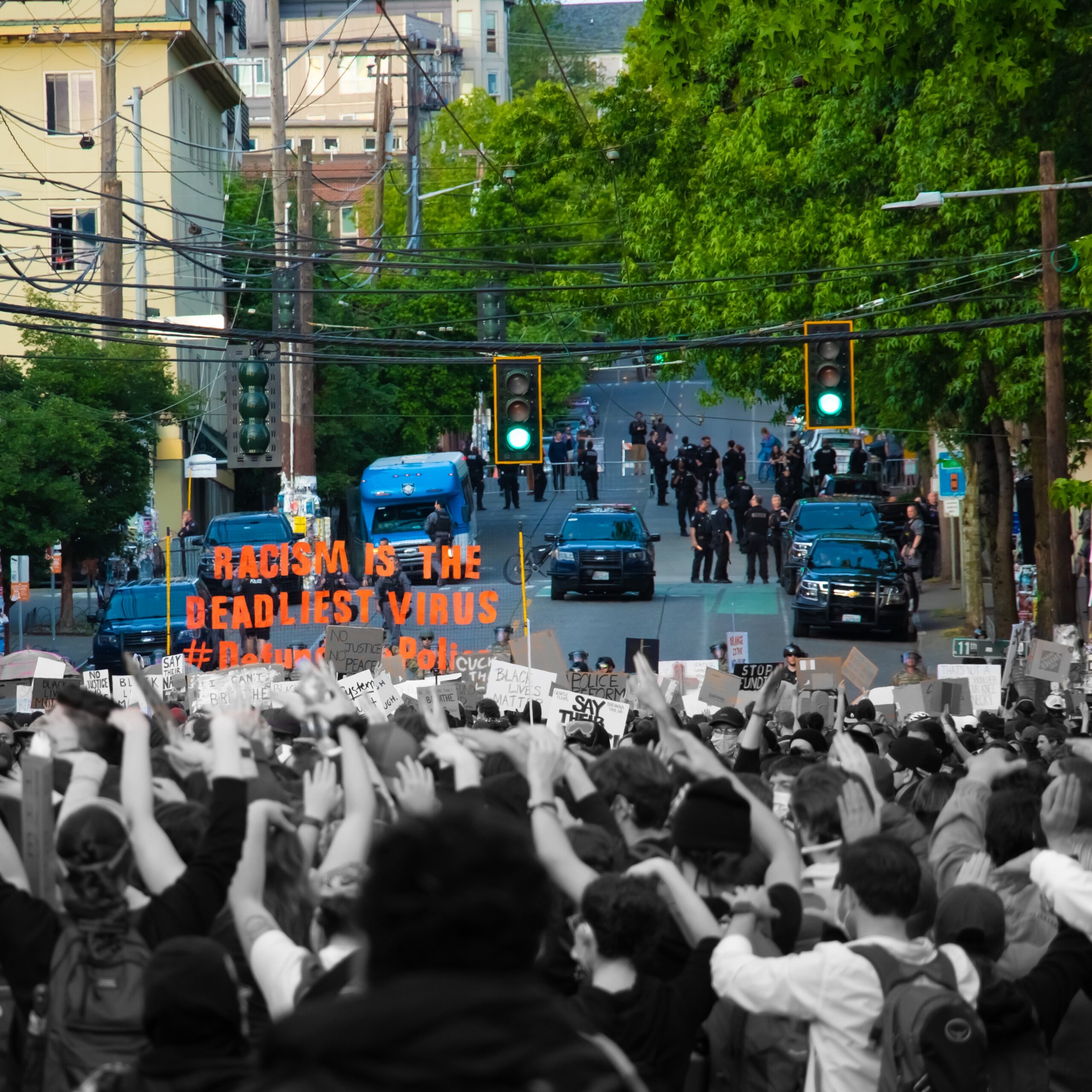 Protesters gather in Seattle, carrying signs and banners. In the distance a group of Seattle PD officers and cars are seen blocking the road. The image is split, with the protesters being in black and white and the police being in color. One banner reads "racism is the deadliest virus"