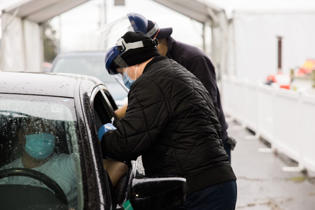 Two males wearing blue surgical masks, clear plastic face shields and black coats stand next to a vehicle. Each male reaches in through a window of the sedan sedan with gloved hands. The manner of the male reaching in to the driver, whose face is largely obscured by a wet windshield, indicates he is administering a shot.