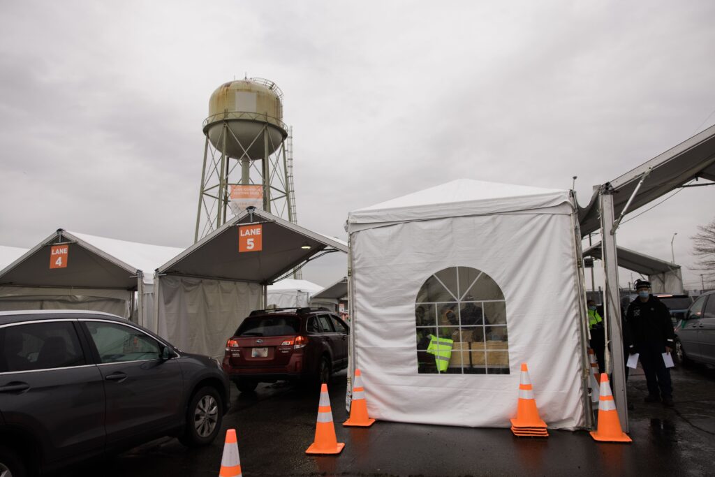 Cars wait in line to pull up inder a white open air tent. Orange traffic cones create a lane for the vehicles. An enclosed tent to the right of the carport houses individuals waiting to serve the vehicles.
